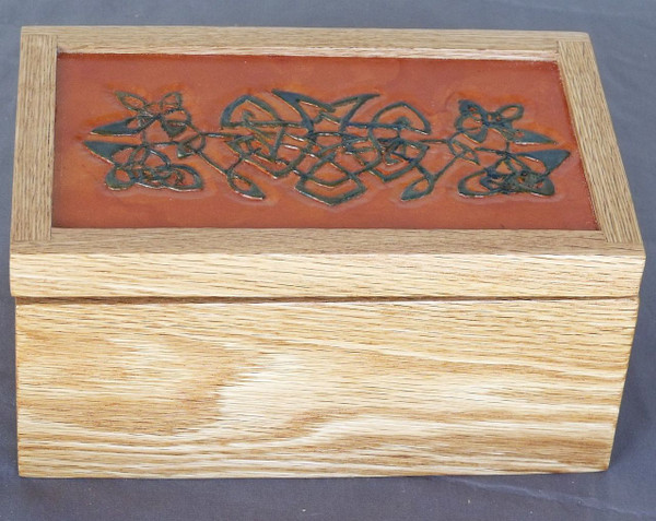 A rectangular red oak keepsake box showcasing a prominent top inset paneled with genuine leather, which is hand-carved with a classic Celtic knot design and dyed deep blue.