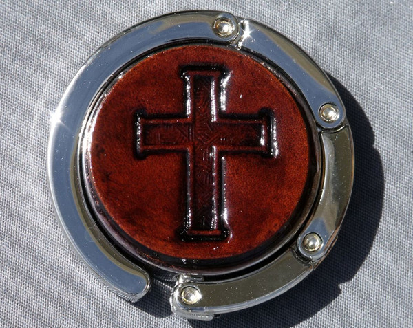 A purse hook with a leather medallion on it.  The medallion has a dark brown background with a cross in the middle. The hook part is wrapped around the center and held closed with small magnets.