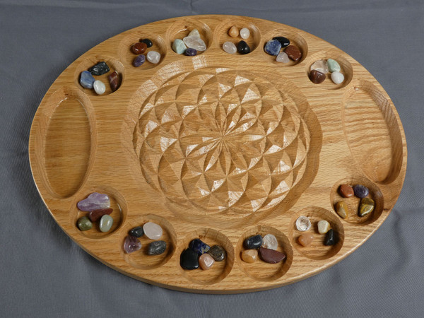 The large deluxe mancala board in oak with the flower of life in the center and beautiful stones in their starting positions.