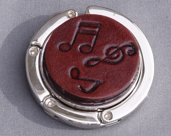 A purse hook with a leather medallion on it.  The medallion has a dark mahogany background with musical notes and symbols randomly placed in the middle. The hook part is wrapped around the center and held closed with small magnets.