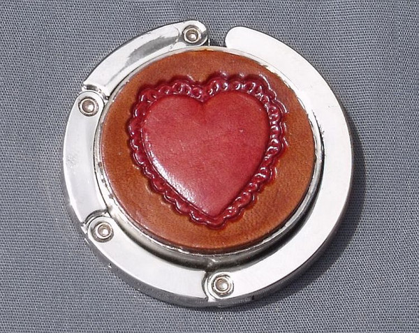 A purse hook with a leather medallion on it.  The medallion has a brown background with a red heart in the middle. The hook part is wrapped around the center and held closed with small magnets.