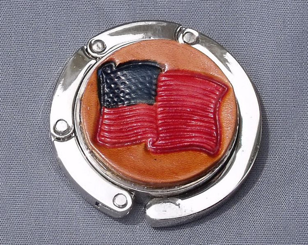 A purse hook with a leather medallion on it.  The medallion has a tan or light brown background with an American flag in the middle. The hook part is wrapped around the center and held closed with small magnets.