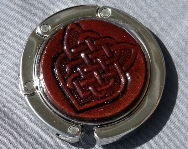 A purse hook with a leather medallion on it.  The medallion has a dark brown background with a celtic knot in the middle. The hook part is wrapped around the center and held closed with small magnets.