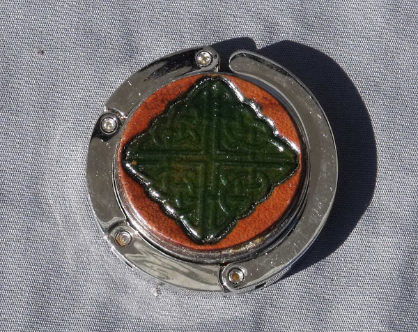 A purse hook with a leather medallion on it.  The medallion has a tan or light brown background with a green celtic knot in the middle. The hook part is wrapped around the center and held closed with small magnets.