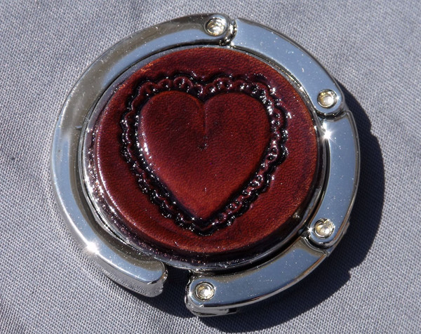 A purse hook with a leather medallion on it.  The medallion has a brown background with a heart in the middle. The hook part is wrapped around the center and held closed with small magnets.