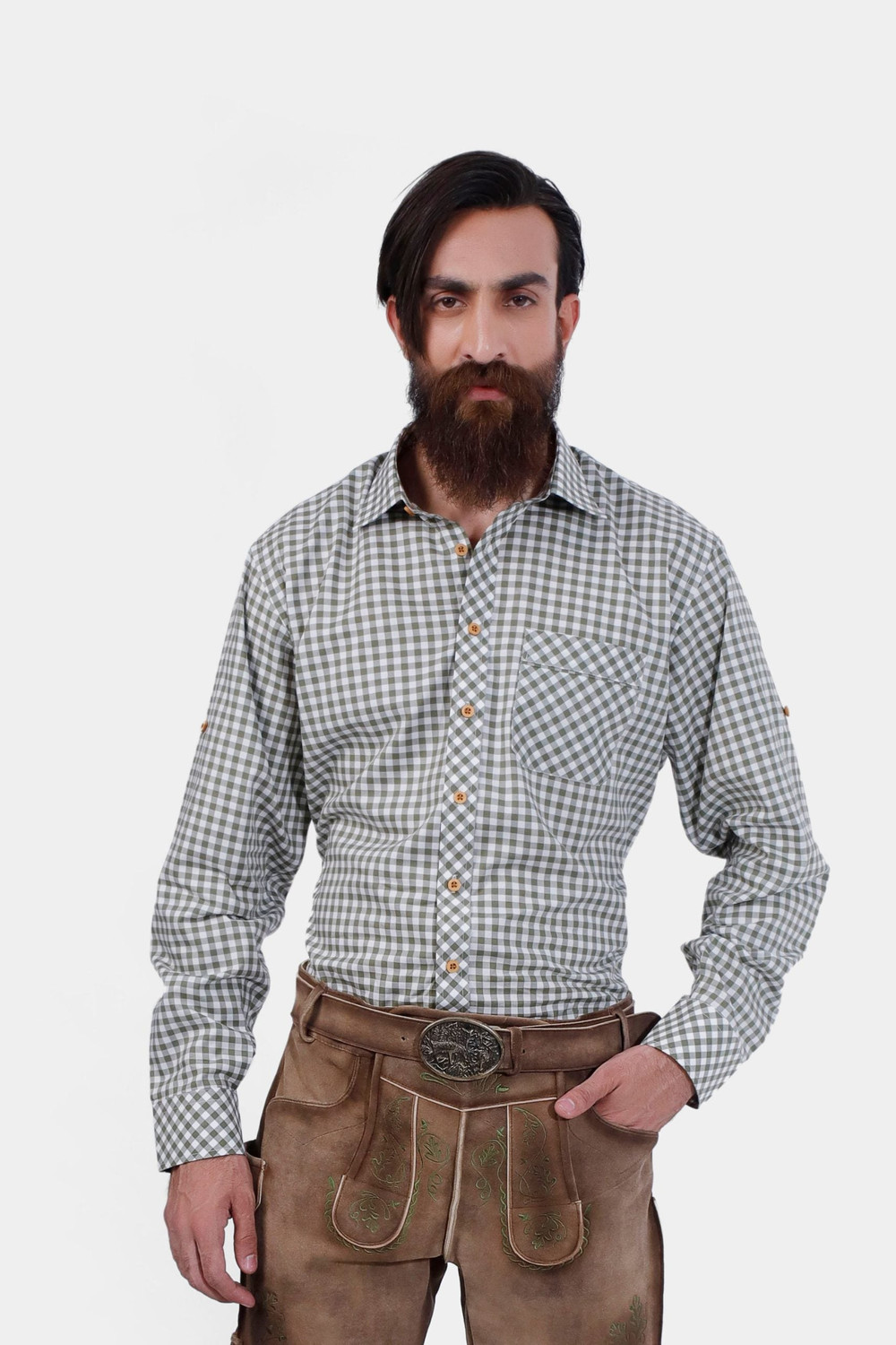 A bearded man wearing the André Lederhosen Shirt, featuring a green and white checkered pattern. He is paired with traditional beige lederhosen embroidered with green detailing. The man stands confidently with one hand in his pocket against a plain white 