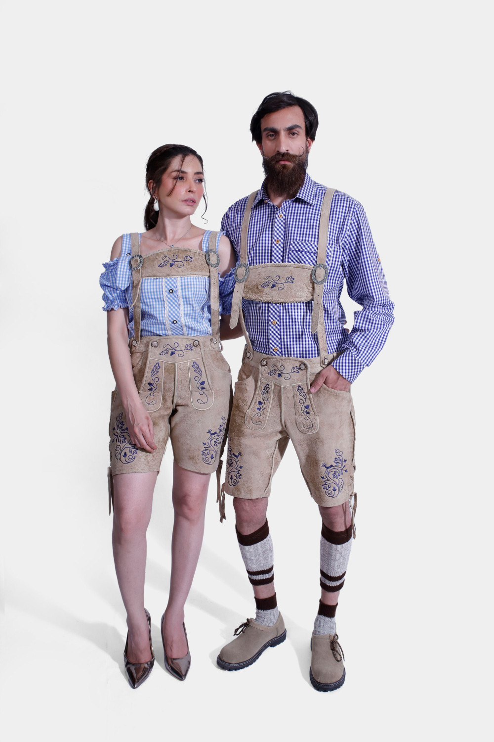 A couple dressed in matching Berliner Couple Outfits. The woman is wearing a blue checkered off-shoulder blouse paired with light tan leather shorts featuring intricate blue embroidery. The man is wearing a blue checkered shirt with light tan leather shor