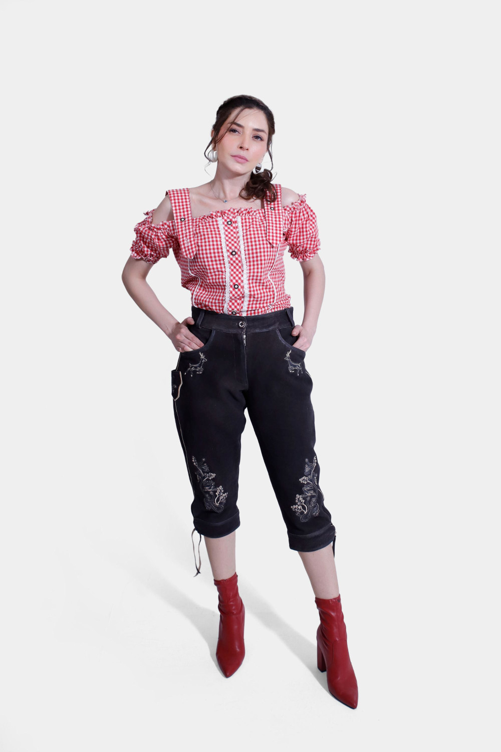 Woman in stylish red and white checkered off-shoulder blouse and black Midnight Raven Lederhosen with intricate silver embroidery, standing confidently. damen lederhose schwarz​, lederhose damen schwarz​