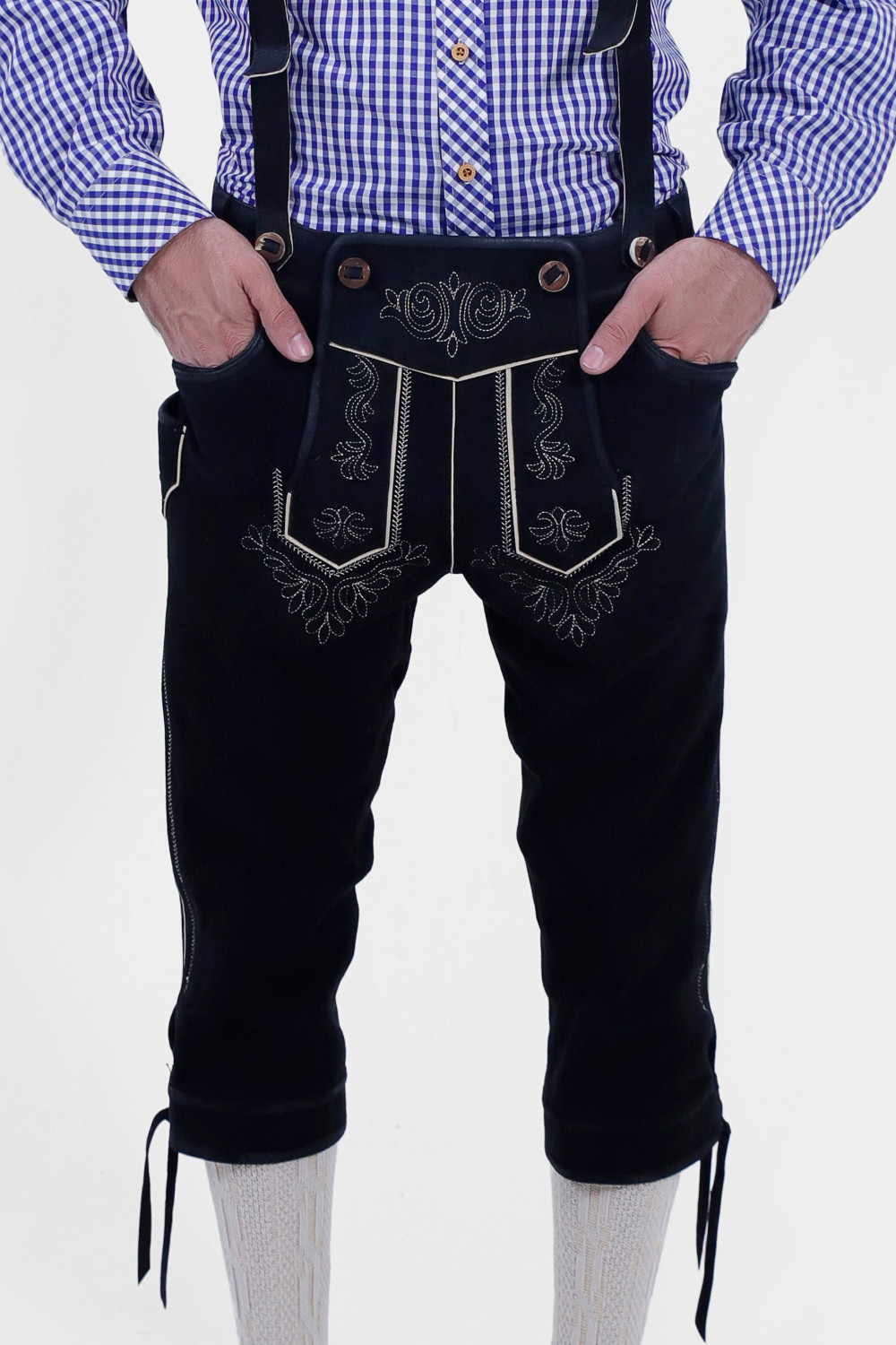 The image shows a pair of traditional black leather Marx Bundhosen with white embroidery, worn with a blue and white checkered shirt. The Bundhosen have intricate designs on the front and sides, and the person has their hands in the front pockets.