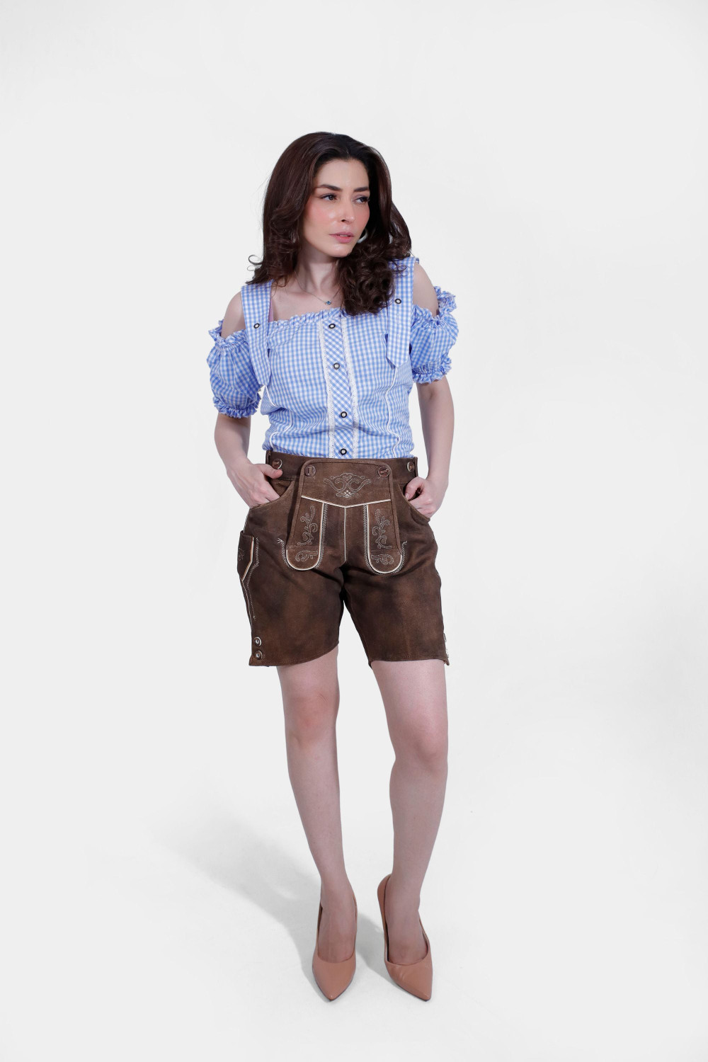 Woman in a blue and white checkered off-shoulder blouse and brown leather lederhosen, standing with hands in pockets, showing front view. damen lederhose tracht​