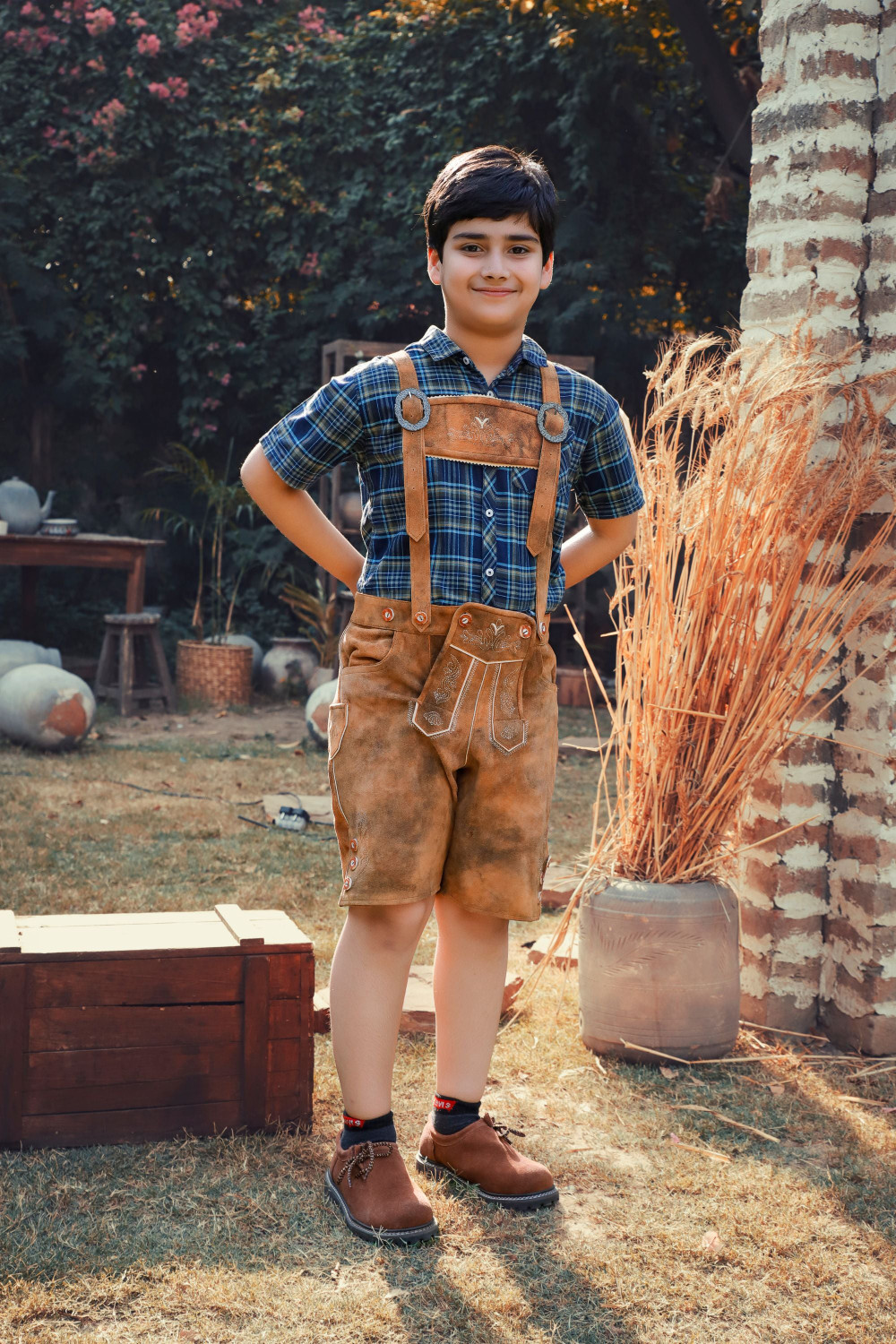 A young boy stands outdoors wearing Jakob Jäger Lederhosen, featuring a rustic amber color and traditional Bavarian design, with suspenders and detailed embroidery. He pairs the lederhosen with a blue plaid shirt and brown shoes, posing confidently with hands on his hips in a charming, rustic garden setting. kinderlederhose
