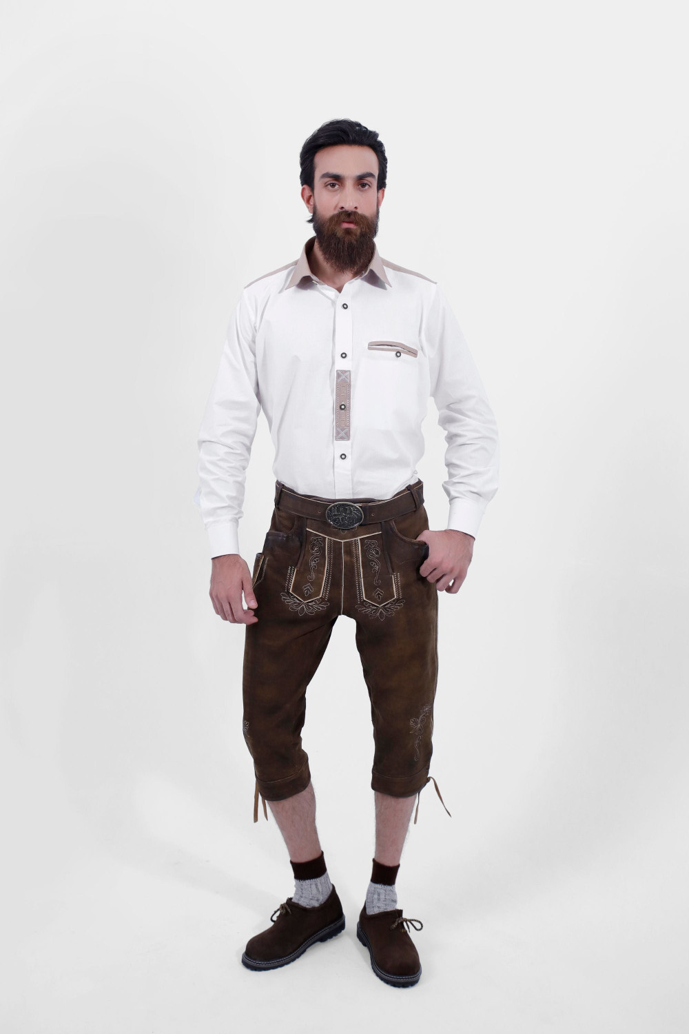 A male wearing a white shirt and Schweinsteiger Bundhosen Outfit features intricate embroidery on the front, a decorative belt buckle, and mid-length. The model's hands are by his sides, and he is wearing brown shoes with grey socks. trachten outfit herren modern​ , trachten outfit herren​