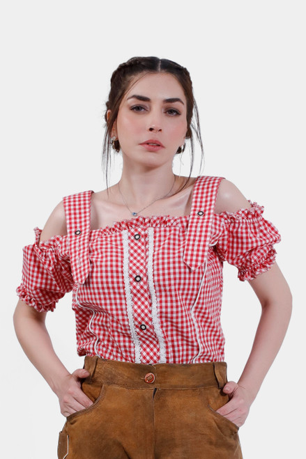 A woman wearing a red checkered off-shoulder traditional blouse with ruffled sleeves and decorative buttons. She pairs the blouse with tan leather lederhosen, standing confidently with her hands in her pockets. Her hair is styled in a braided updo, giving