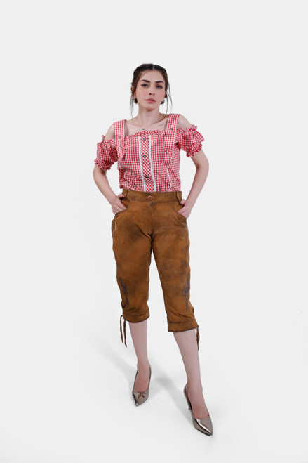 The woman is standing in a similar pose as the first image but slightly turned to the side. She is wearing a red and white checkered, off-shoulder blouse with ruffled sleeves, and the "Kirsten Dunst Lederhosen" brown leather pants. Her hands are in her pockets, and she is looking directly at the camera. lederhose knielang damen​
