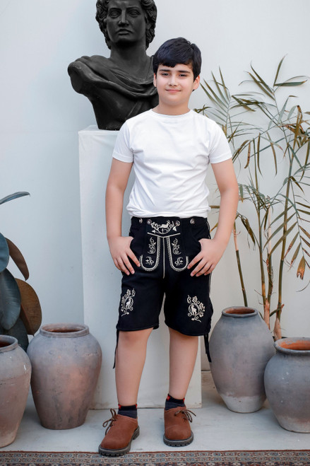 A child standing with hands in pockets, wearing Rilke Lederhosen in Midnight Edelweiss color, detailed with intricate white embroidery, and posing in front of a statue and plants. lederhose kinder​ , Lederhosen für Kleinkinder 