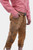 Close-up side view of a man wearing a red and white checkered shirt paired with brown leather Bundhosen. The image focuses on the detailed embroidery on the side of the pants and the rugged texture of the leather. His hand rests in the pocket, showcasing 