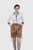 A woman wearing a white button-down shirt with a polka-dotted collar and cuffs, paired with tan Lederhosen shorts featuring intricate floral embroidery. The shorts are secured with a decorative belt with an ornate buckle. She stands confidently with her h