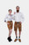 A couple dressed in matching Neuschwanstein Couple Outfits. The woman is wearing a crisp white blouse paired with golden-brown leather shorts featuring intricate embroidery and black lace-up boots. The man is wearing a white button-down shirt with golden-
