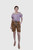 A woman wearing a purple and white checkered off-shoulder blouse with puffed sleeves, paired with tan Lederhosen shorts featuring floral embroidery. She is wearing a wide belt with an ornate buckle and beige high heels, standing confidently with one hand 