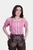 A woman is wearing a pink and white checkered off-shoulder blouse with button details, paired with brown embroidered lederhosen shorts. Her hands are casually placed in the pockets, and her hair is styled in loose waves. The lederhosen feature intricate f