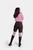 The woman is standing with her body slightly turned, showing the back of her outfit. The brown lederhosen shorts and the back of the pink and white checkered blouse with a zipper detail are visible. She is wearing knee-high brown boots, and her hair is st