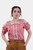 A woman wearing a red checkered off-shoulder traditional blouse with ruffled sleeves and decorative buttons. She pairs the blouse with tan leather lederhosen, standing confidently with her hands in her pockets. Her hair is styled in a braided updo, giving