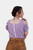 Woman wearing the Rihanna Traditional Blouse in purple checkered pattern, back view. The blouse features off-the-shoulder sleeves with ruffled edges, lace detailing along the back seam, and is paired with traditional leather trousers. The intricate design