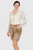 A woman wearing a cream-colored blouse with lace sleeves paired with beige Lederhosen shorts. The shorts feature decorative buttons on the sides and intricate embroidery. She stands confidently with one hand in her pocket