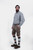A man with a beard is standing in a three-quarter view against a white background, wearing a grey and white checkered shirt and grey leather lederhosen. He has his right hand in the pocket of the lederhosen, and is also wearing green and grey knee-high so