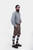 A man with a beard is standing in a three-quarter view against a white background, wearing a grey and white checkered shirt and grey leather lederhosen. He has his right hand in the pocket of the lederhosen, and is also wearing green and grey knee-high so