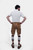 A man with a beard is standing with his back to the camera, hands on hips, wearing a short-sleeved white shirt and brown leather lederhosen. He is also wearing brown and grey knee-high socks and brown shoes. The lederhosen has pockets on both sides and is