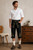A man wearing traditional Bavarian black embroidered kniebundhose with gold and white floral details, a white button-down shirt, white socks, and brown leather shoes, standing in a rustic room with a wooden chest and chair.