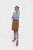 A woman is standing in a Clara Lederhosen skirt with her back to the camera, showing the rear view of the skirt and her blue and white checkered off-shoulder blouse, with her hair braided and tied back.