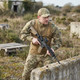 Man wearing Kombat UK - UBACS Tactical Fleece Ripstop Top in British Terrain Pattern Camouflage crouching outdoors with rifle