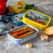 The image captures a delightful and colorful gourmet setting with two tins of sardines. One tin is open, revealing sardines covered in a vibrant red sauce, possibly tomato-based, with visible herbs, which suggests a flavorful preparation. The tin of sardines rests on a textured surface, next to a dark blue napkin and a silver fork, ready for serving.

Behind the open tin, there's a closed tin with a bright yellow lid, featuring a whimsical illustration of sardines driving a classic car, enhancing the playful nature of the product branding. The closed tin's design suggests a connection to a fun, lively theme, possibly a nod to the product's origin or the flavors within.

The setting includes natural elements like flowers and a red bell pepper to the side, contributing to the fresh and natural appeal of the meal. Additionally, two golden-brown biscuits are placed casually near the open tin, suggesting a pairing for the sardines that adds a textural contrast with a slight sweetness or buttery flavor to complement the savory fish.

The entire composition is lively and suggests a casual, yet thoughtful dining experience. The variety of textures and colors from the food to the tableware creates a visually appealing scene that invites the viewer to enjoy a taste of these sardines, which seem to promise both quality and flavor.