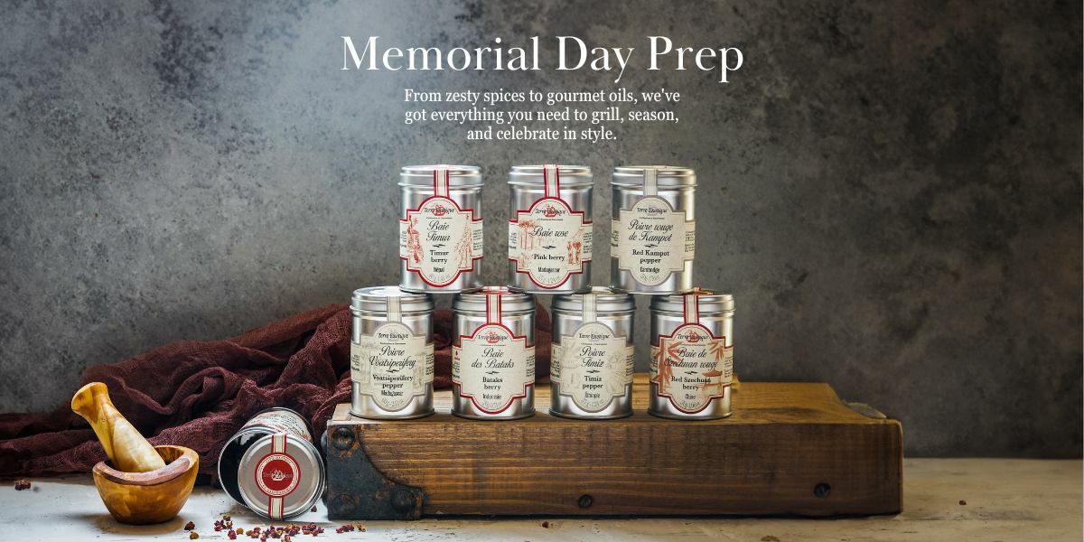 This image features an assortment of spice tins artfully arranged on a wooden cutting board, which is placed on a table. The spice tins have a vintage design, with labels indicating various flavors such as "Fine Thyme," "Pink Peppercorn," "Basilic," "Red Kampot Pepper," among others. A mortar and pestle with some spices in it, along with a scattered few pink peppercorns, are also visible on the table, suggesting the preparation of fresh spices. A deep red cloth adds a warm touch to the composition. The background is a textured grey surface that complements the rustic aesthetic. At the top of the image, the text "Memorial Day Prep" is prominently displayed, followed by a message that reads, "From zesty spices to gourmet oils, we've got everything you need to grill, season, and celebrate in style." The overall mood is one of high-quality culinary preparation for Memorial Day festivities.