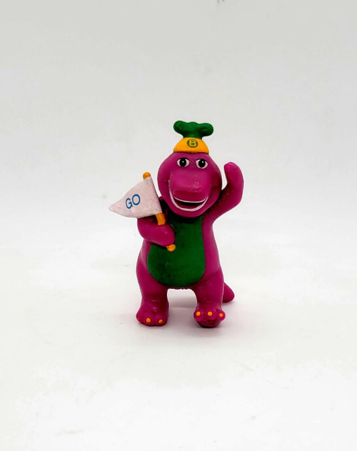 Barney & Friends PVC Cheerleader Barney Toy Figure, colorful pink dinosaur mascot in cheerleading outfit with a green top, yellow hat with a letter B, holding a small white flag with 'GO' written on it, and waving one arm.