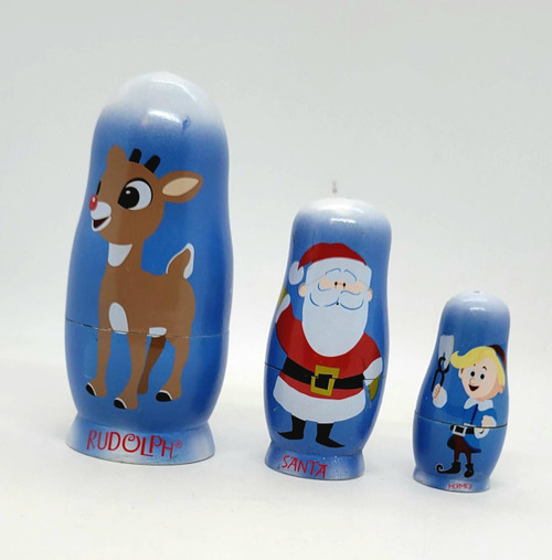 Babooshkah Rudolph The Red-Nosed Reindeer 50th Anniversary Wood Nesting Doll Set, set of three blue nesting dolls featuring Rudolph the Red-Nosed Reindeer, Santa Claus, and Hermey, decorated with festive holiday illustrations.