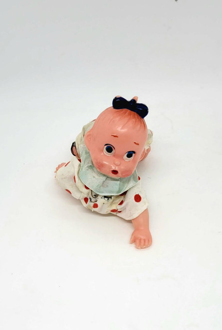 Unknown Vintage Mechanical Wind-up Crawling Baby Toy, old plastic baby doll with a black bow on its head, dressed in a white polka-dot outfit with a green bib, lying on a white background.
