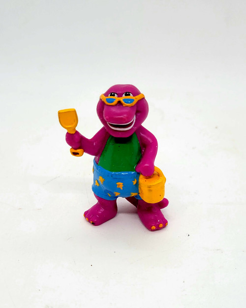1993 Barney & Friends PVC toy figure of Barney at the beach.  The figure stands 2.5 inches tall and features Barney wearing blue shorts with yellow stars.  Barney is also carrying a yellow bucket and shovel.