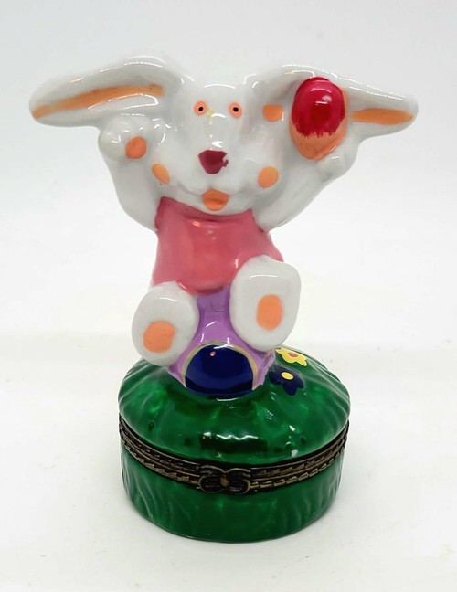 Unknown Easter Bunny Hinged Trinket Box, ceramic bunny figurine with a smiling face, pink outfit, sitting on a green grassy base with colorful flowers, used for storing small keepsakes or jewelry.