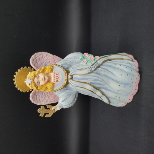 Novelino 1993 Marjorie Sarnal Angels of the Month - June, a decorative angel figurine with golden curly hair, a white and pink gown with gold accents, pink wings with a textured pattern, a golden crown, and a gold cross in hand, standing against a black background.