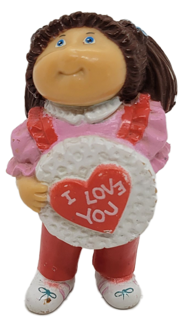 Cabbage Patch Kids 1984 PVC figure of a brunette hair girl wearing pink shirt and red overalls and white shoes by OAA.  The girl is holding a heart with "I Love You" with white ribbon in front of her and white flowers held behind her back.  The figure stands 2.25-inches tall.