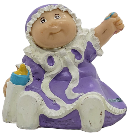 Cabbage Patch Kids 1984 PVC figure of a baby girl wearing purple dress and hat with white trim and white shoes by OAA.  The girl is holding a bottle in her right hand and left hand raised.  The figure stands 1.75-inches tall.