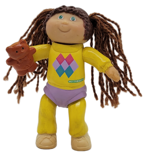 Cabbage Patch Kids 1984 poseable figure of a brown yarn hair girl wearing yellow shirt with multicolor diamonds, yellow tights, purple shorts and tan shoes by OAA.  The girl is holding a teddy bear in her right hand. The figure stands 3-inches tall.
