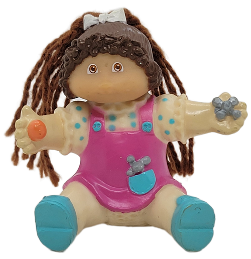 Cabbage Patch Kids 1985 figure of a brown yarn hair girl wearing purple overalls over pink shirt with blue spots by OAA.  The girl is sitting down with ball in right hand and jacks in her left hand. The figure stands 2.5-inches tall.