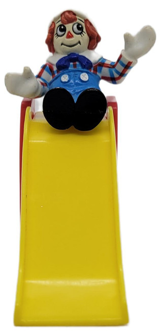 MacMillan Inc. McDonald's Happy Meal Toy 1989 Raggedy Ann & Andy Playground Raggedy Andy With A Slide, toy featuring Raggedy Andy sitting on a yellow slide, waving with a cheerful expression.