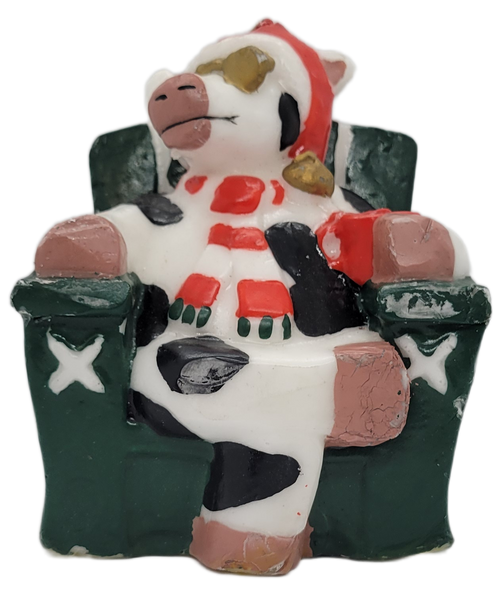 Novelty Christmas candle of a Holstein cow wearing Santa hat and red and white stripped scarf while sitting in a green chair. The cow is also wearing gold sunglasses and holding a red mug in arms. The single wick candle is by Boston Warehouse Trading Corp and was made in China.