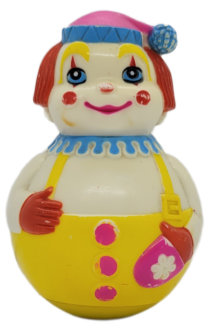 The First Years 1972 Roly Poly Clown toy by Kidde Products Inc. The clown is wearing yellow pants with purple buttons and pocket and red gloves. The clown is wearing a pink hat with blue trim and blue collar. As the clown is moved, the clown makes a metal clinking sound.