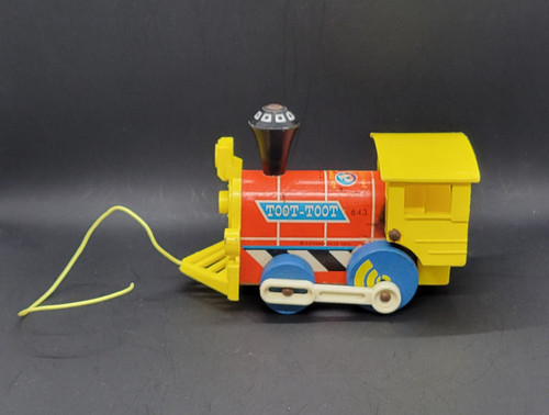 Vintage Fisher-Price 1964 #643 Toot-Toot Engine pull along toy. Wooden cut-out train engine with a rounded boiler that has red paper lithographs on the sides with a blue "sign" marked "Toot Toot" in white. The engine has a yellow plastic front grill and cowcatcher, a yellow plastic cab with a roof, a BLACK wooden smoke stack with small white squares on top, and 4 BLUE wooden wheels with white plastic pistons attached. When pulled, the pistons move and the engine makes a "chug-chug" sound. The model number is marked in small print by the cow catcher. Measures 6-1/4" long, 4-1/4" high, 3-1/4" wide.